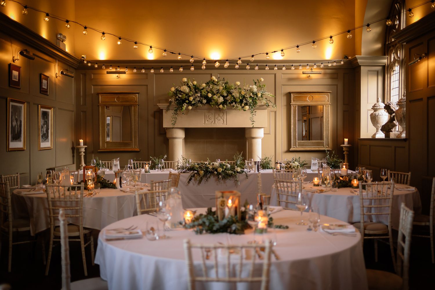 Wedding reception room with romantic string lighting and floral centerpieces on round tables. Ellingham Hall