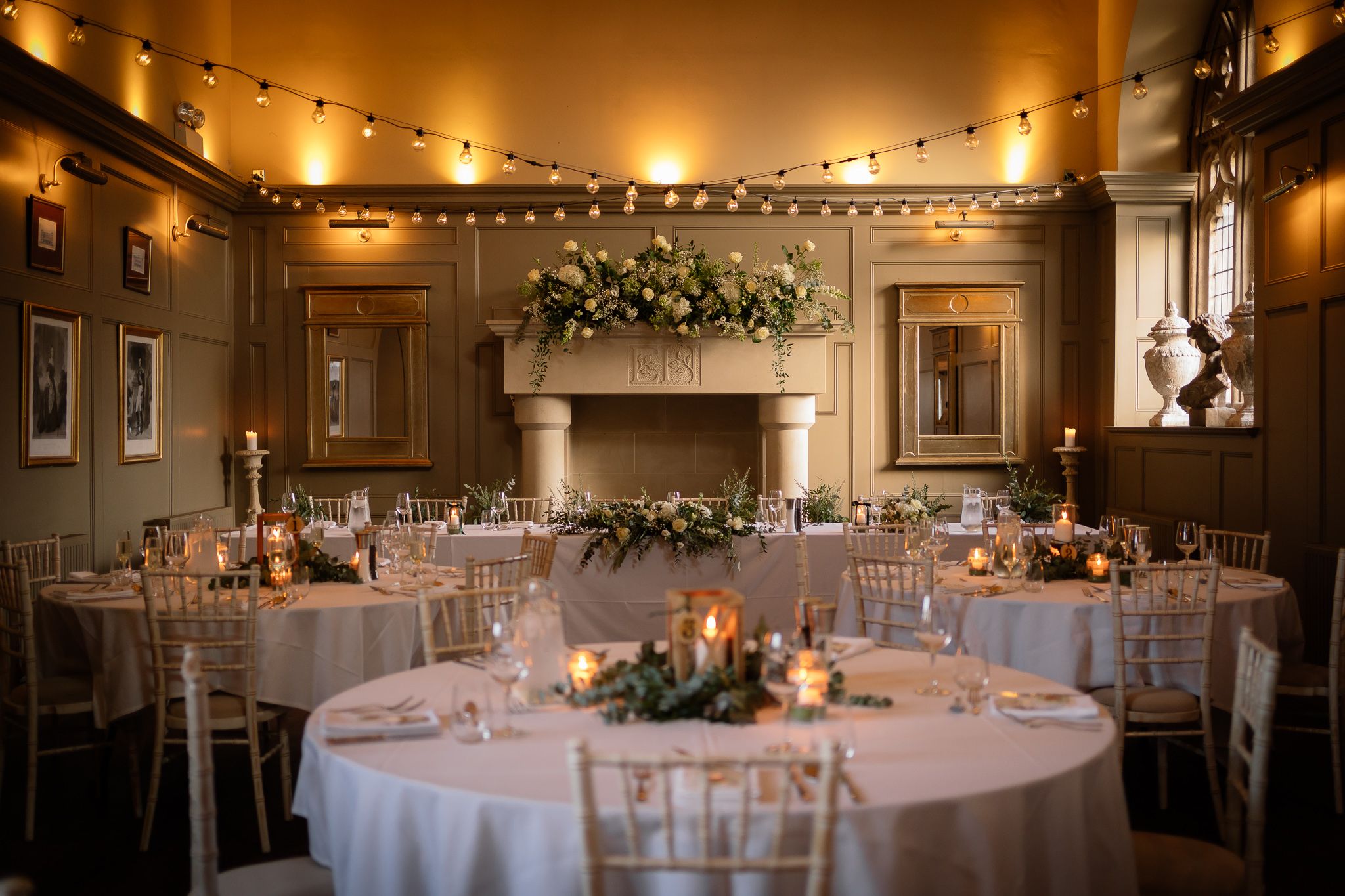 Wedding reception room with romantic string lighting and floral centerpieces on round tables. Ellingham Hall