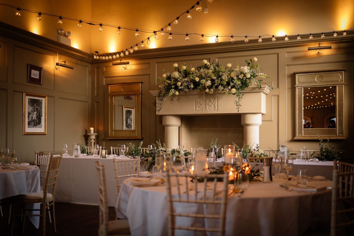 Warm-lit banquet hall with string lights and round tables arranged for an elegant evening event. Ellingham Hall