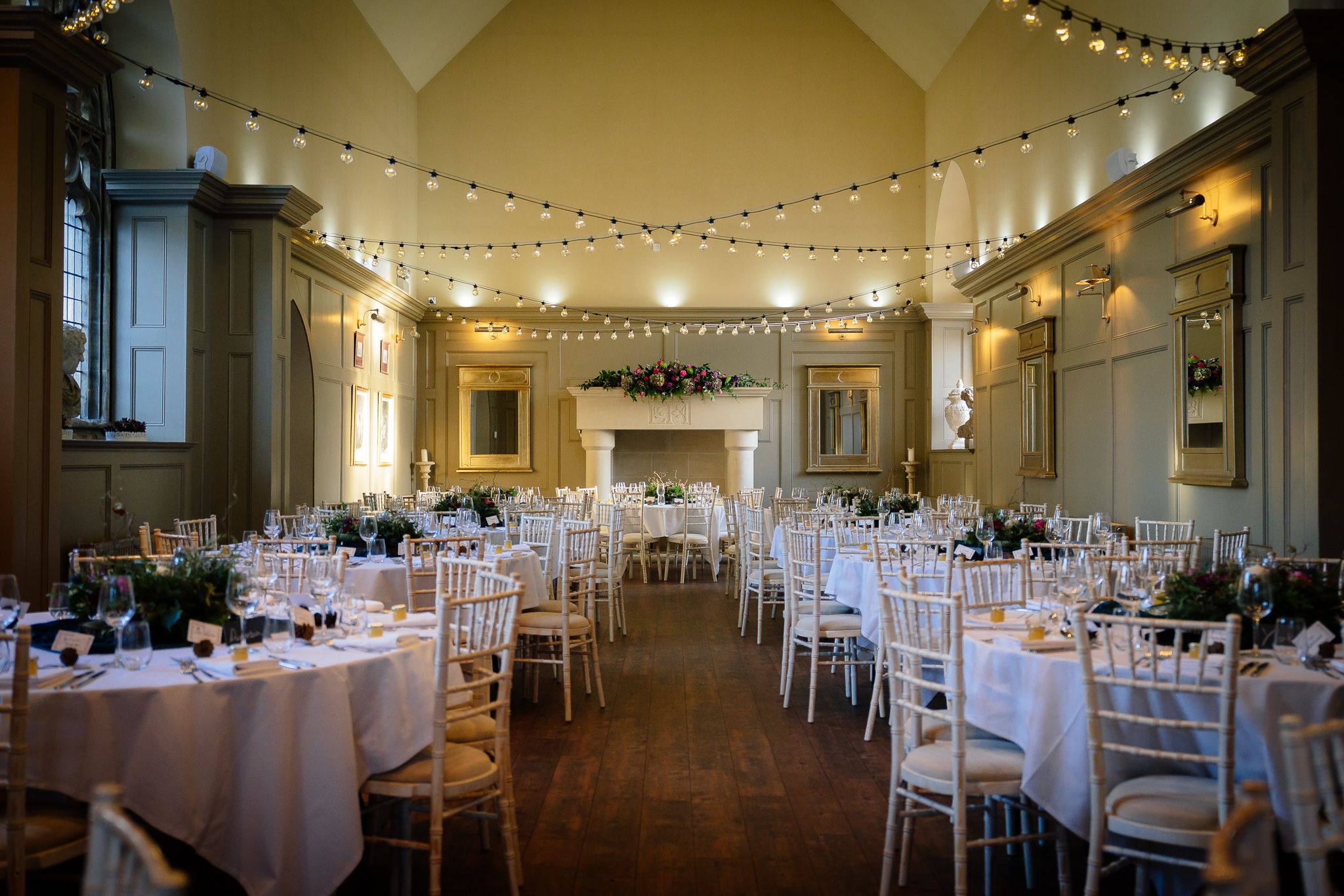 Elegant wedding reception venue with string lights and white tables set for dining in a classic ballroom setting. Ellingham Hall