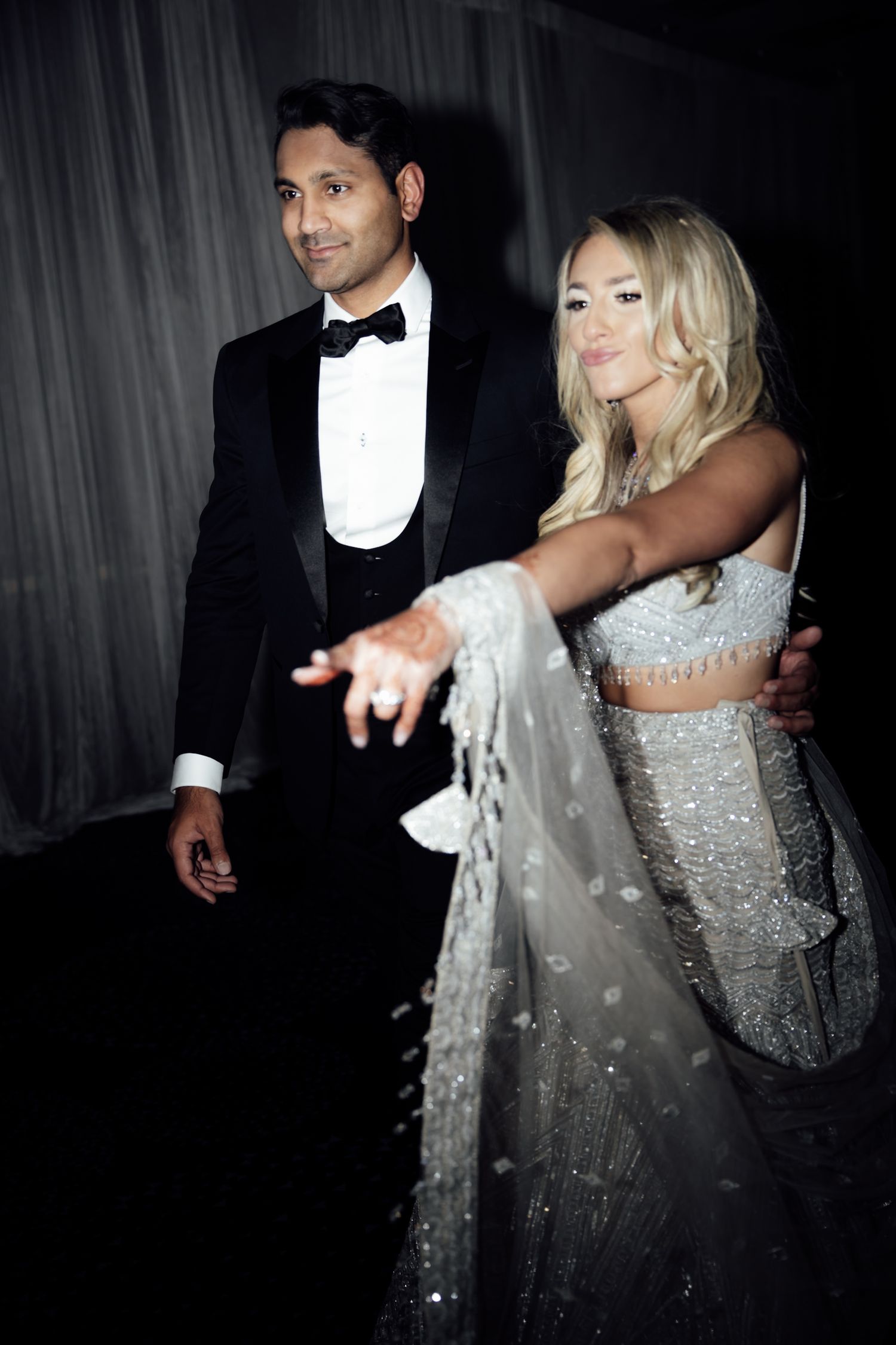 A couple in formal attire dances together, with the woman wearing a sparkly silver dress and the man in a black tuxedo.