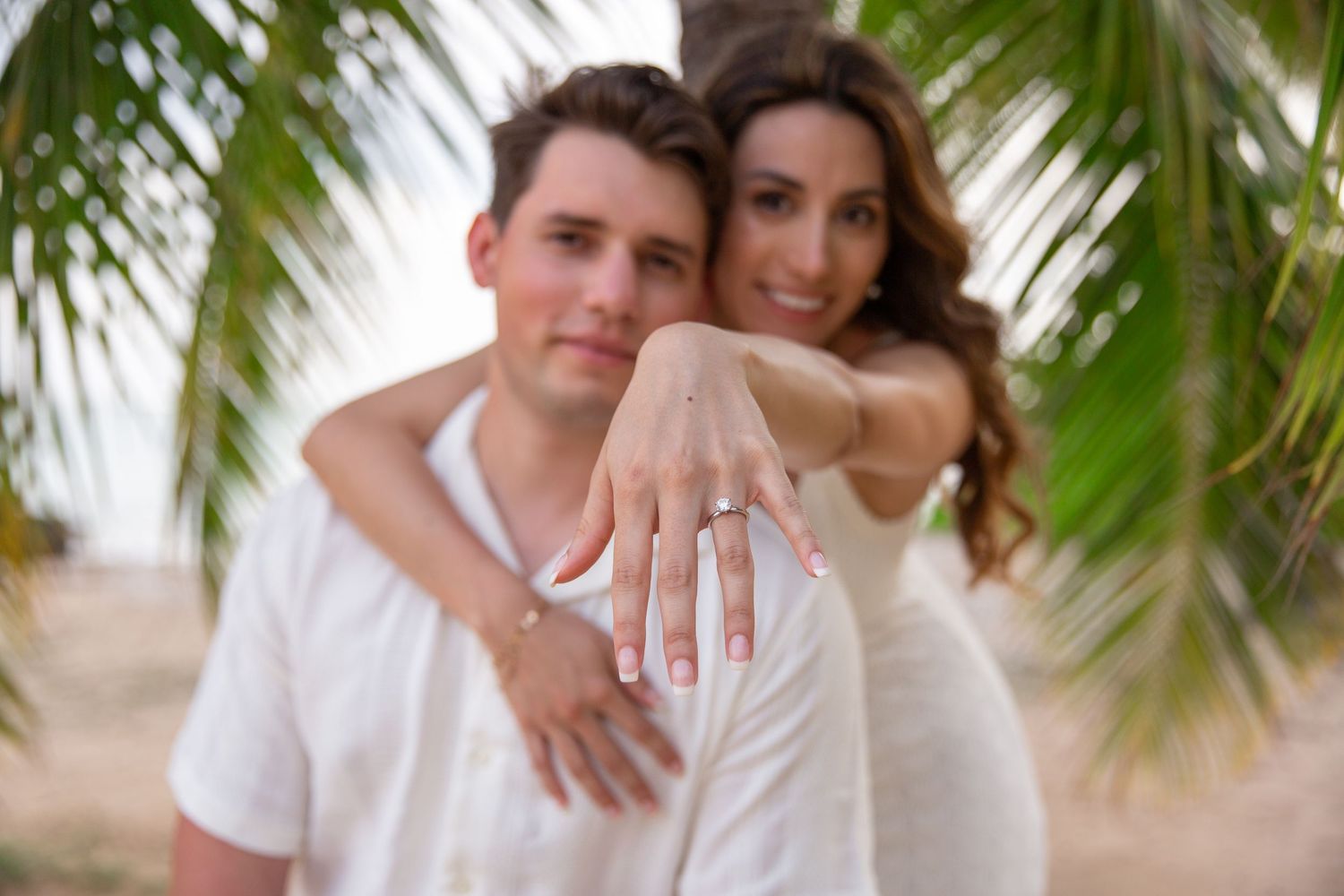 A couple poses together in white outfits on a beach with palm trees at Waialae Beach in Kahala.