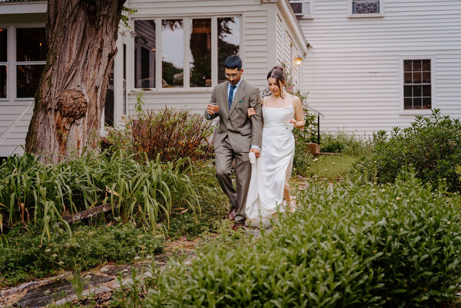 A bride and groom walk together through a lush garden path beside a white farmhouse on their wedding day.