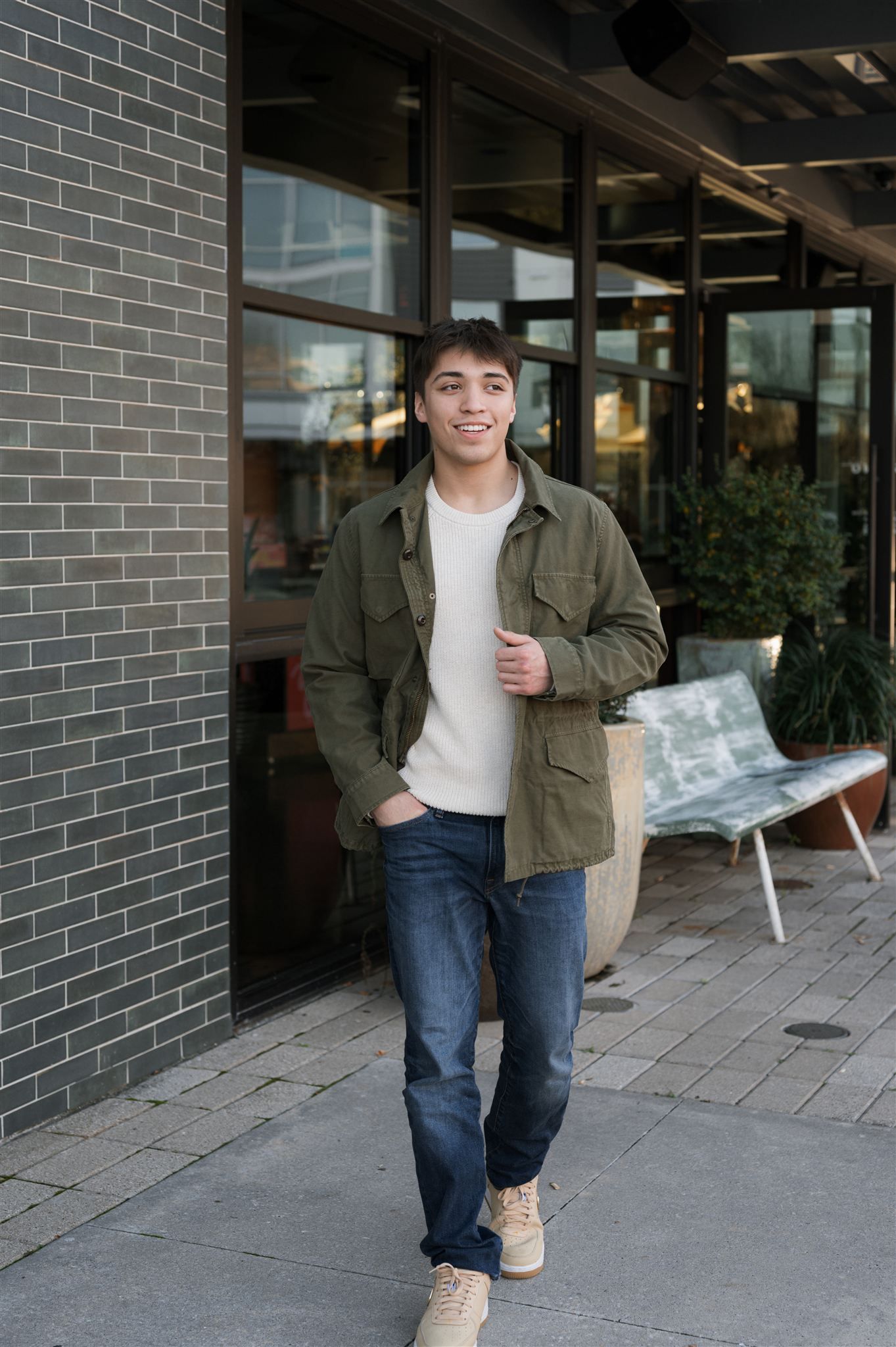 A person in an olive jacket and jeans stands casually outside a brick building with glass windows.