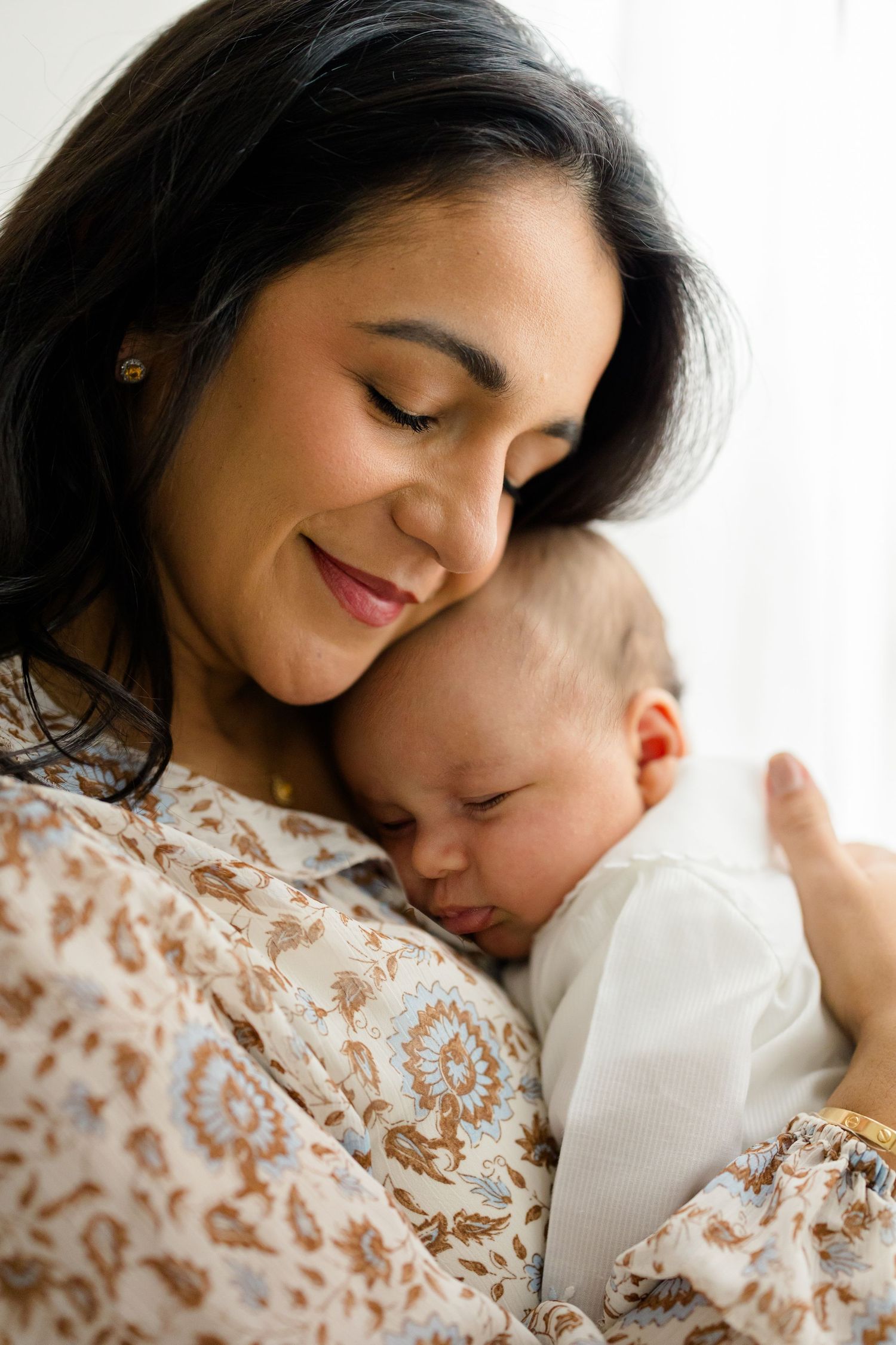 A tender moment captured in soft lighting shows a mother and newborn baby embracing in a white and floral lace outfit.