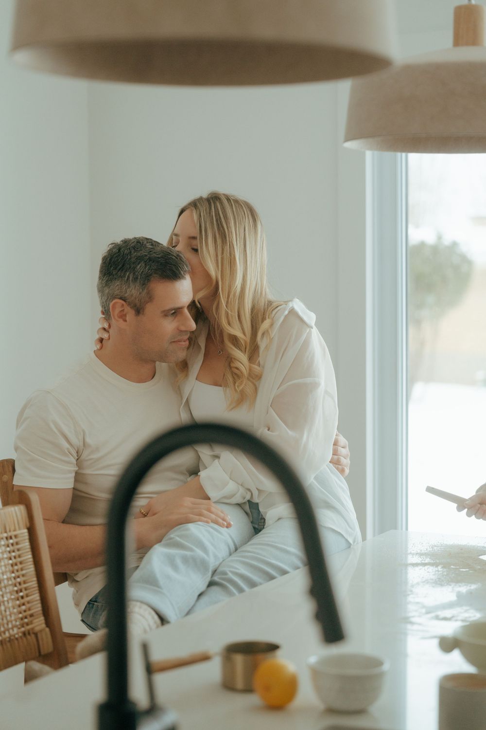 A romantic couple shares an intimate moment in their bright kitchen with a black faucet and citrus on the counter.