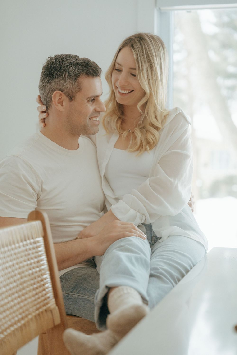 Intimate couple in white clothing sharing a tender moment while sitting on a rattan chair in a bright room.