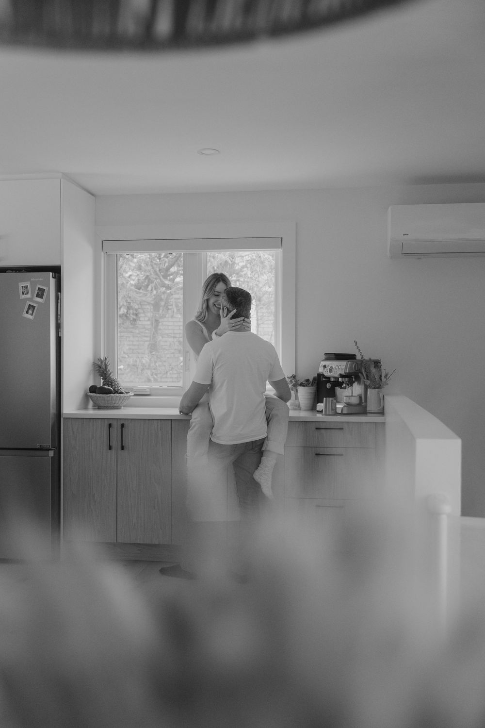 Black and white photo of a couple embracing in a modern kitchen with natural light streaming through the window.