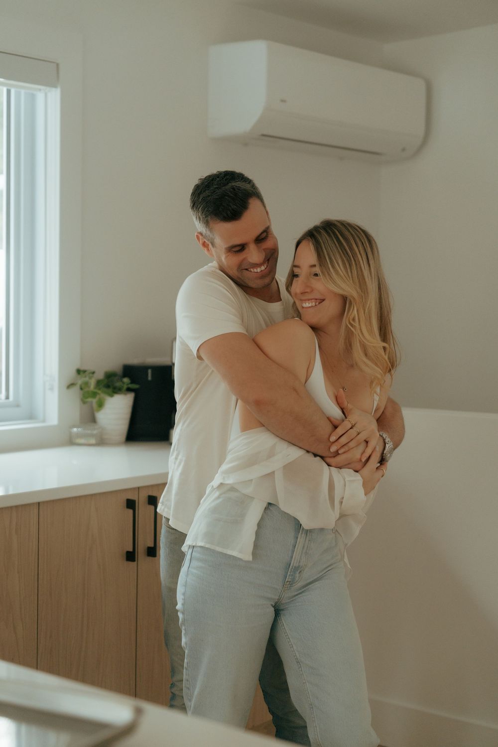A couple embraces and laughs together in a bright, minimalist white room with wooden accents.