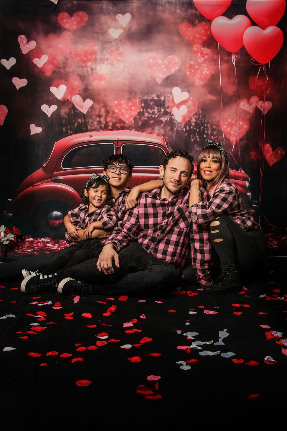 A Valentine's themed photo shoot with red heart balloons, rose petals, and a vintage red car against a dark background.