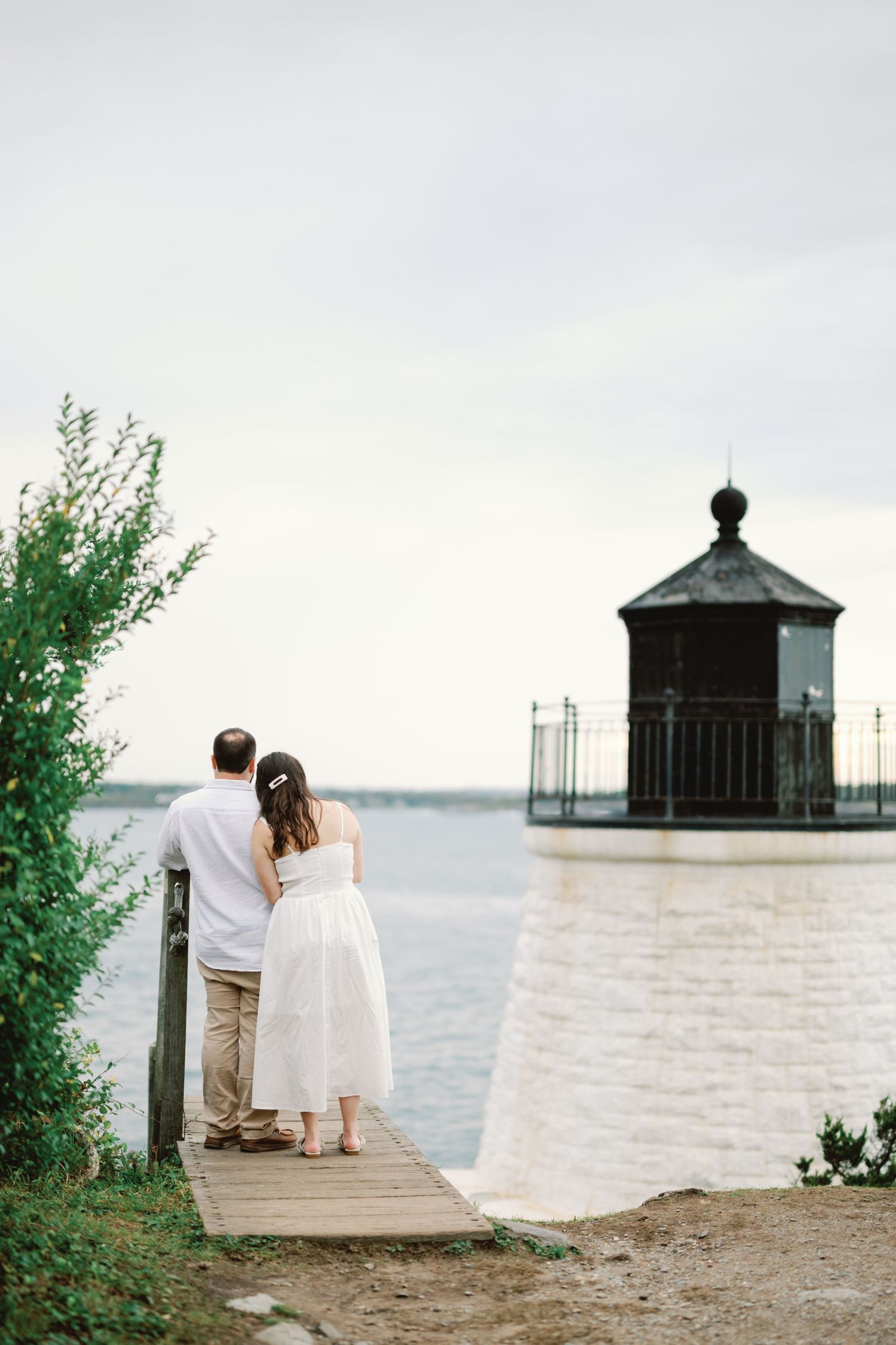 Couple standing on wooden dock overlooking lighthouse and water.