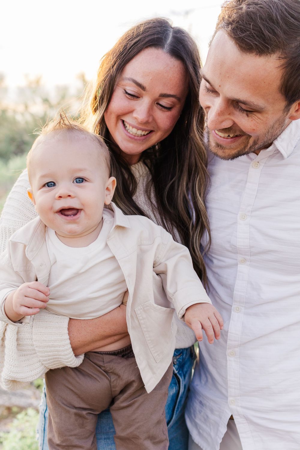 Light and airy family portrait of two parents looking down at infant son smiling in the Mesa desert.
