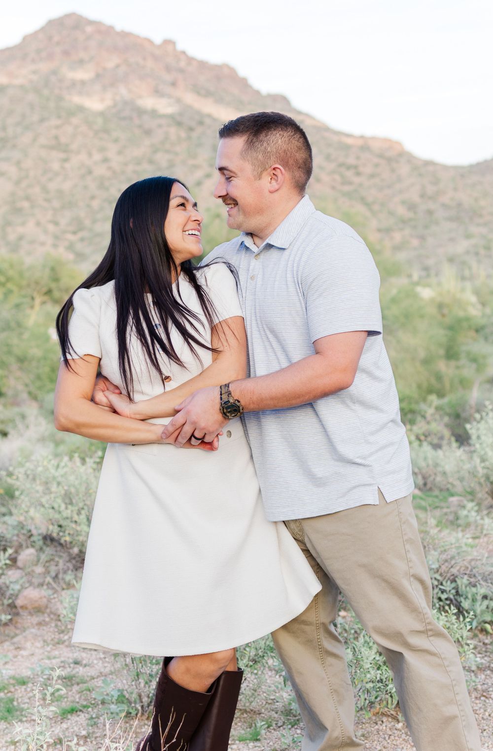 Light and airy portrait of a young married couple side hugging and smiling at each other in front of a mountain in the Arizona desert.
