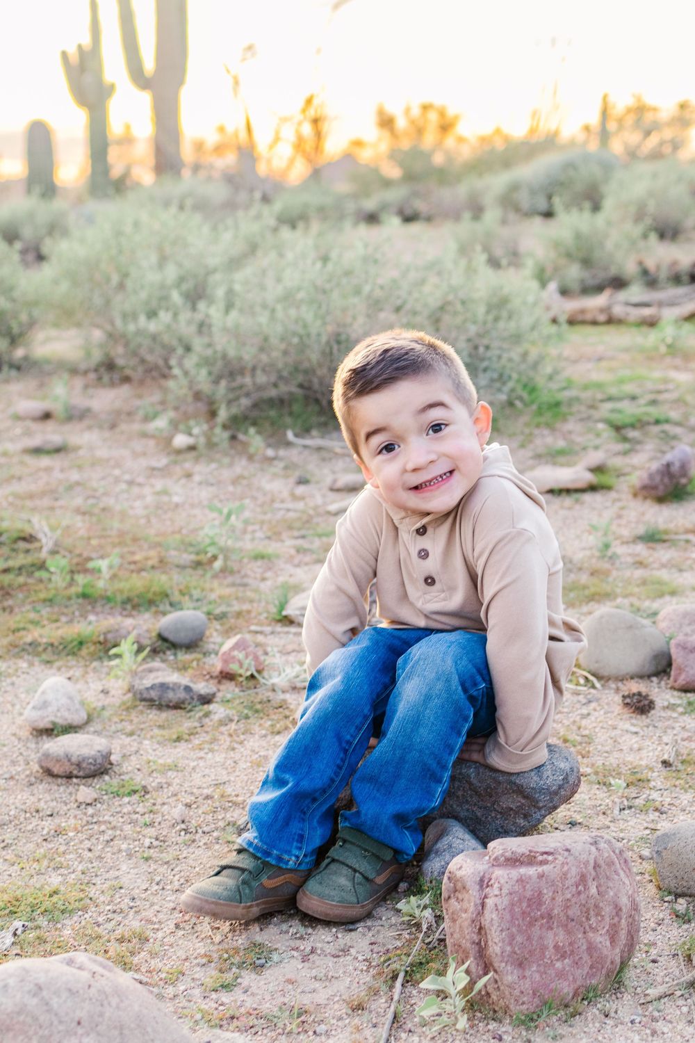 Portrait of young boy smiling and sitting on a rock in the Mesa, Arizona desert during golden hour with his hands placed on the rock.