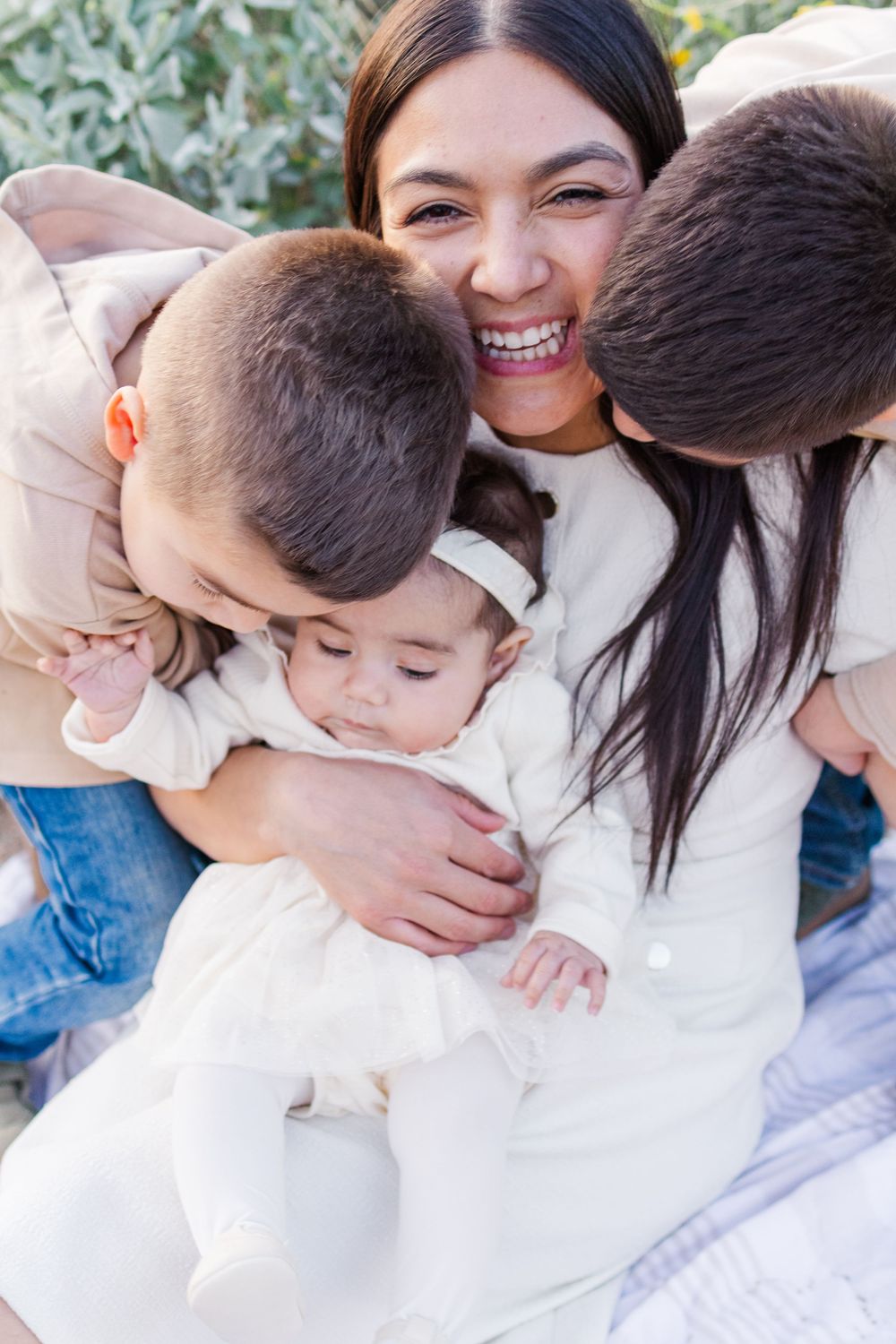 Light and airy portrait of young mom with a group hug from her three young kids sitting on a white blanket in the desert during golden hour.