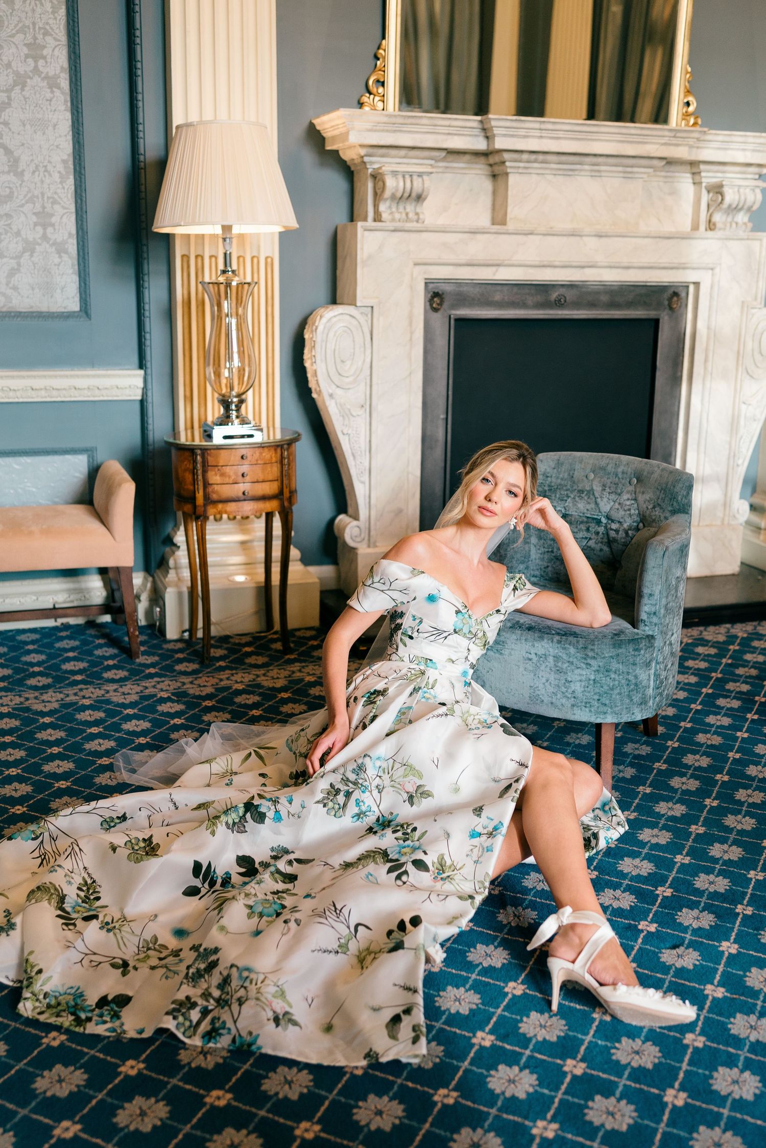 A person in a floral white dress and heels lounges on a blue patterned carpet in an elegant room.