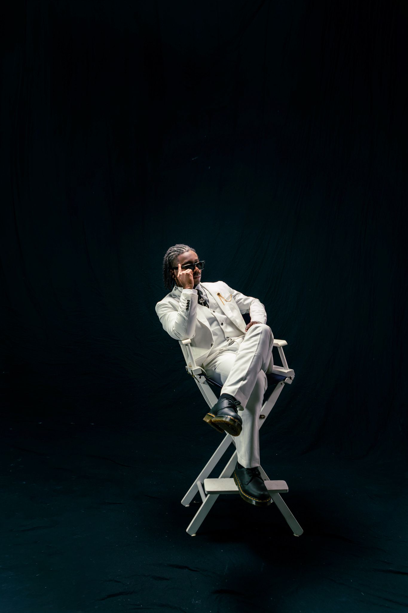 A figure in a white suit sits in a director's chair against a dark background in a dramatic studio photoshoot.