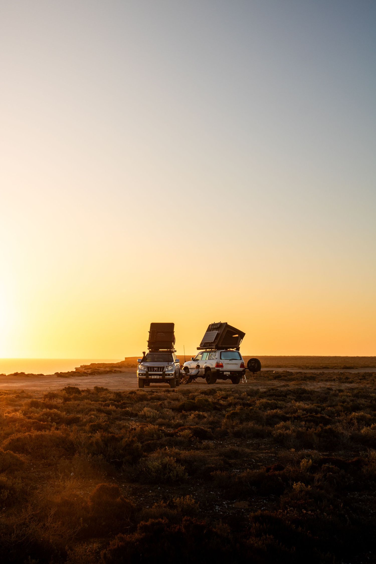 Two 4WD vehicles with rooftop tents parked in open outback landscape at sunset.