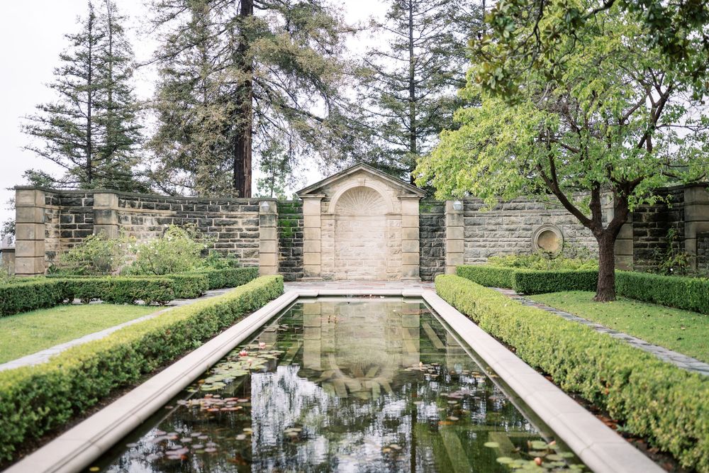 Stone walkway with white balustrades leads through a formal garden with evergreen trees and landscaping.