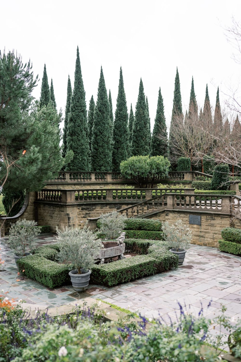 Elegant stone terrace garden features cypress trees, trimmed boxwoods, and flowering plants in a classic Italian design.