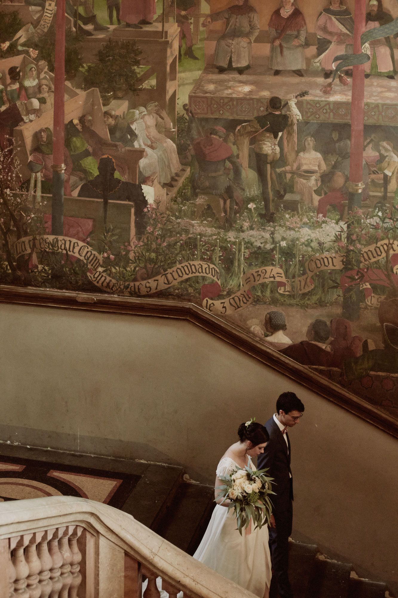 A couple descends an ornate staircase with a vintage mural adorning the wall above.