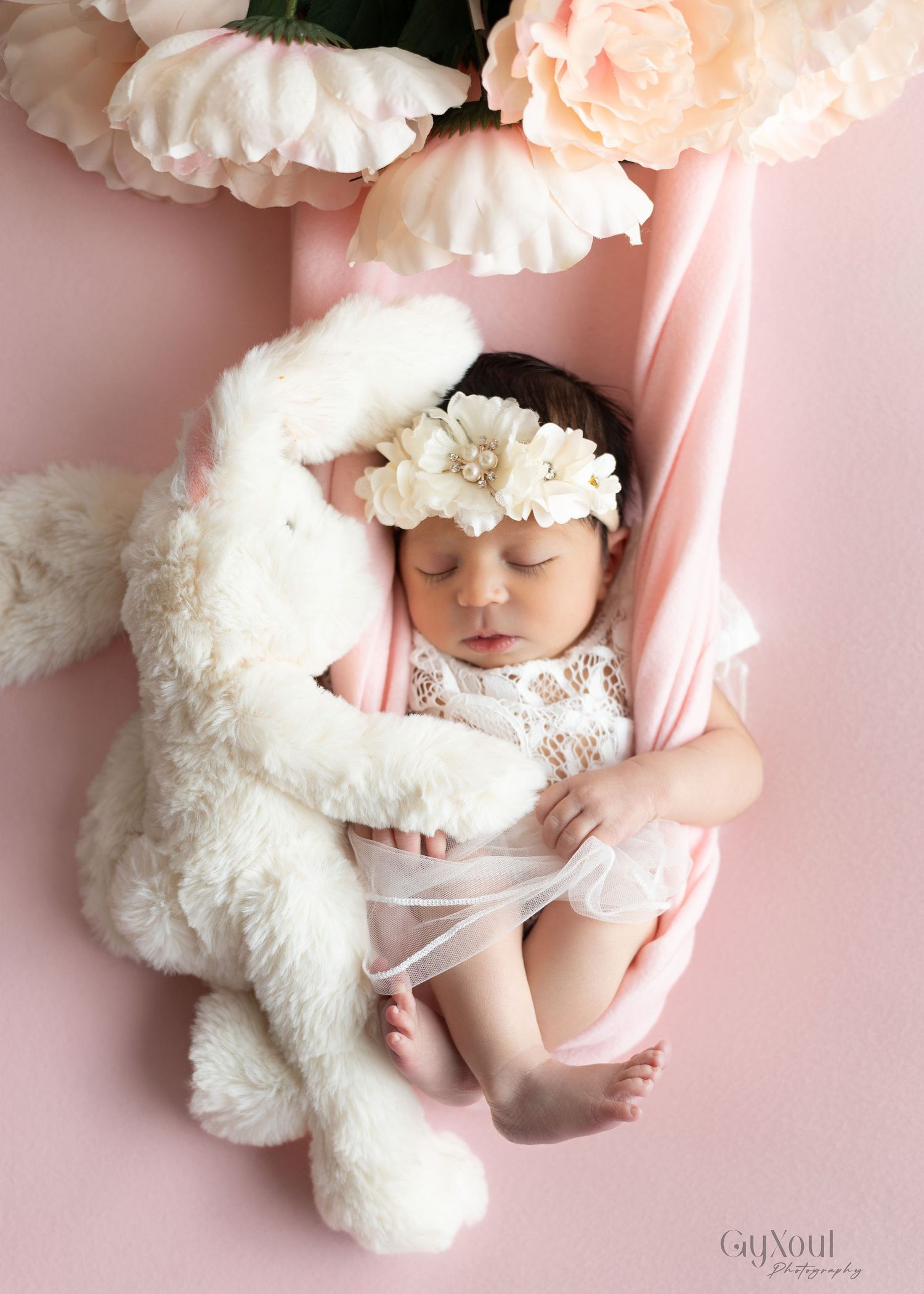 A sleeping baby in white lace with floral headband rests on pink fabric next to a plush white bunny toy.