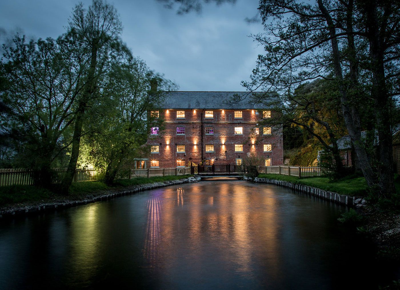 Illuminated brick building reflected in calm water at dusk with trees silhouetted against the evening sky.