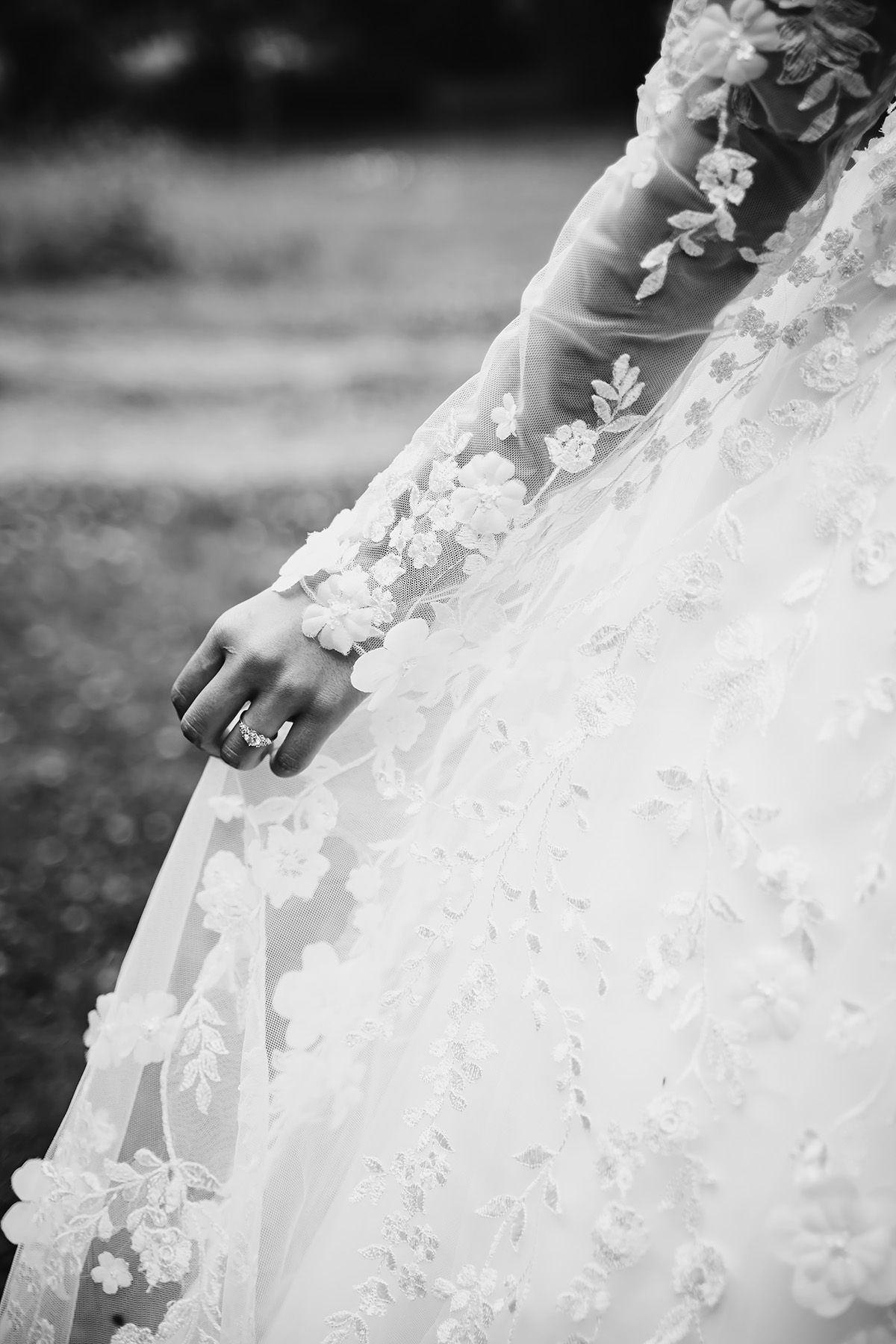 Detaljfoto av en hand som håller upp en brudklänning, armen är klädd i spets och tyget är vitt och skirt med broderade blommor.
