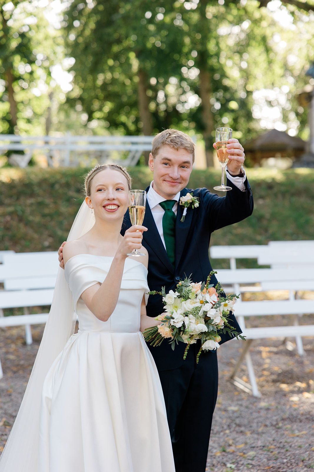 Newlywed couple celebrating with champagne toasts in an outdoor garden setting with white fences and trees.