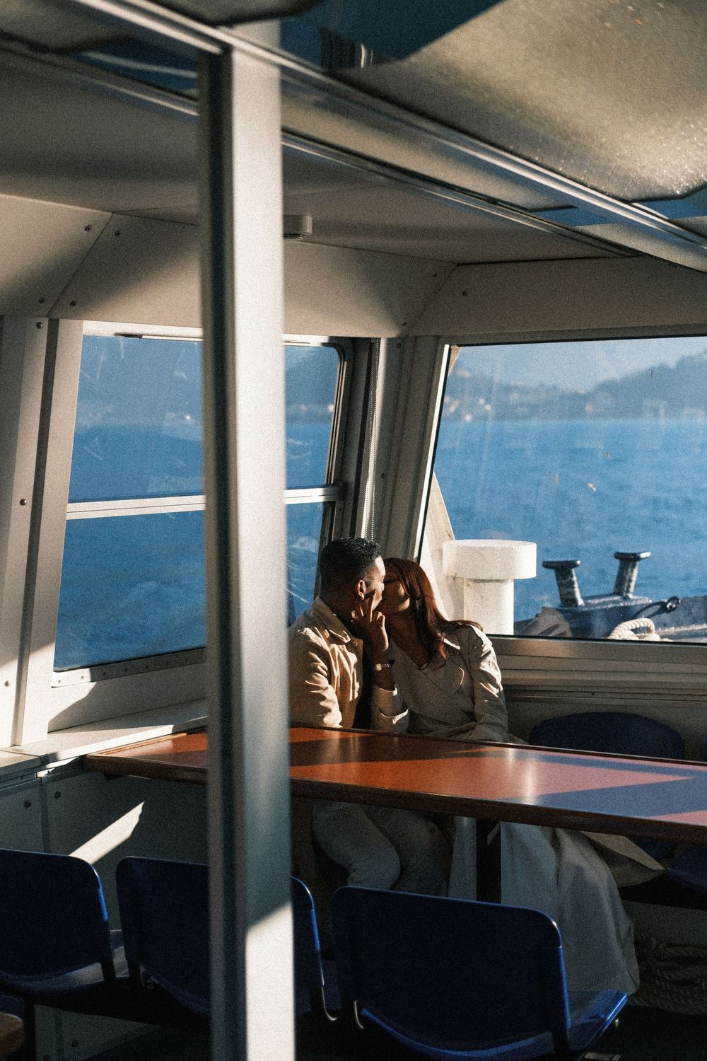 Interior view of a boat cabin with wooden tables, blue chairs and windows overlooking the water on a sunny day.