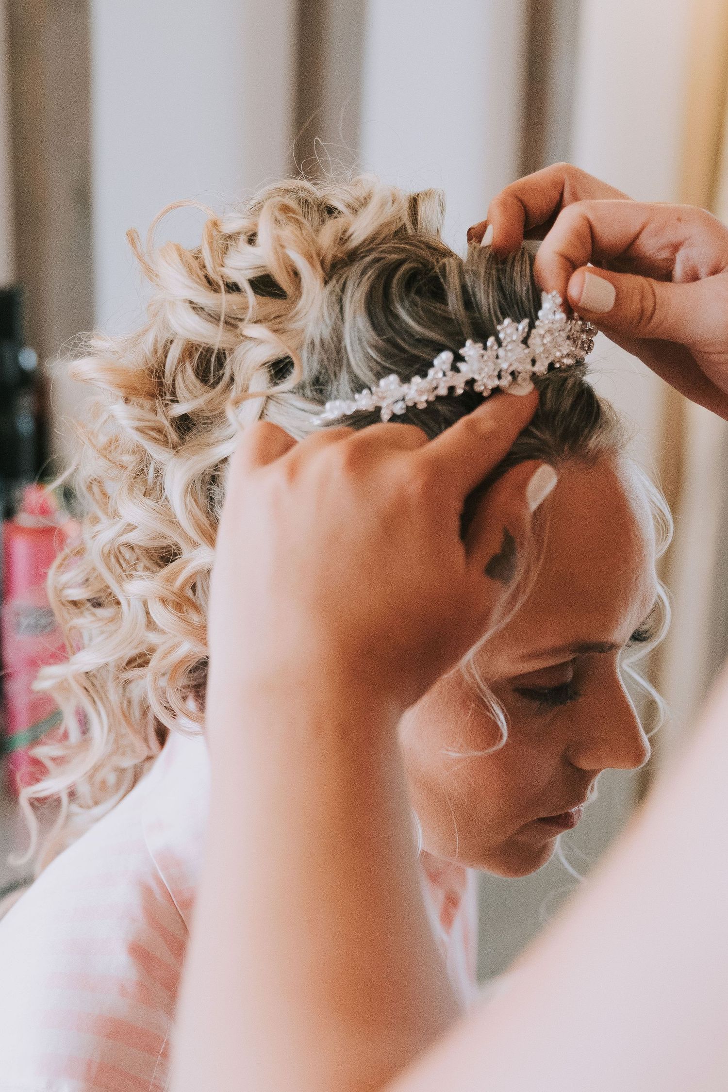 Close-up side view of a delicate pearl and crystal bridal headpiece being placed in blonde curled hair.