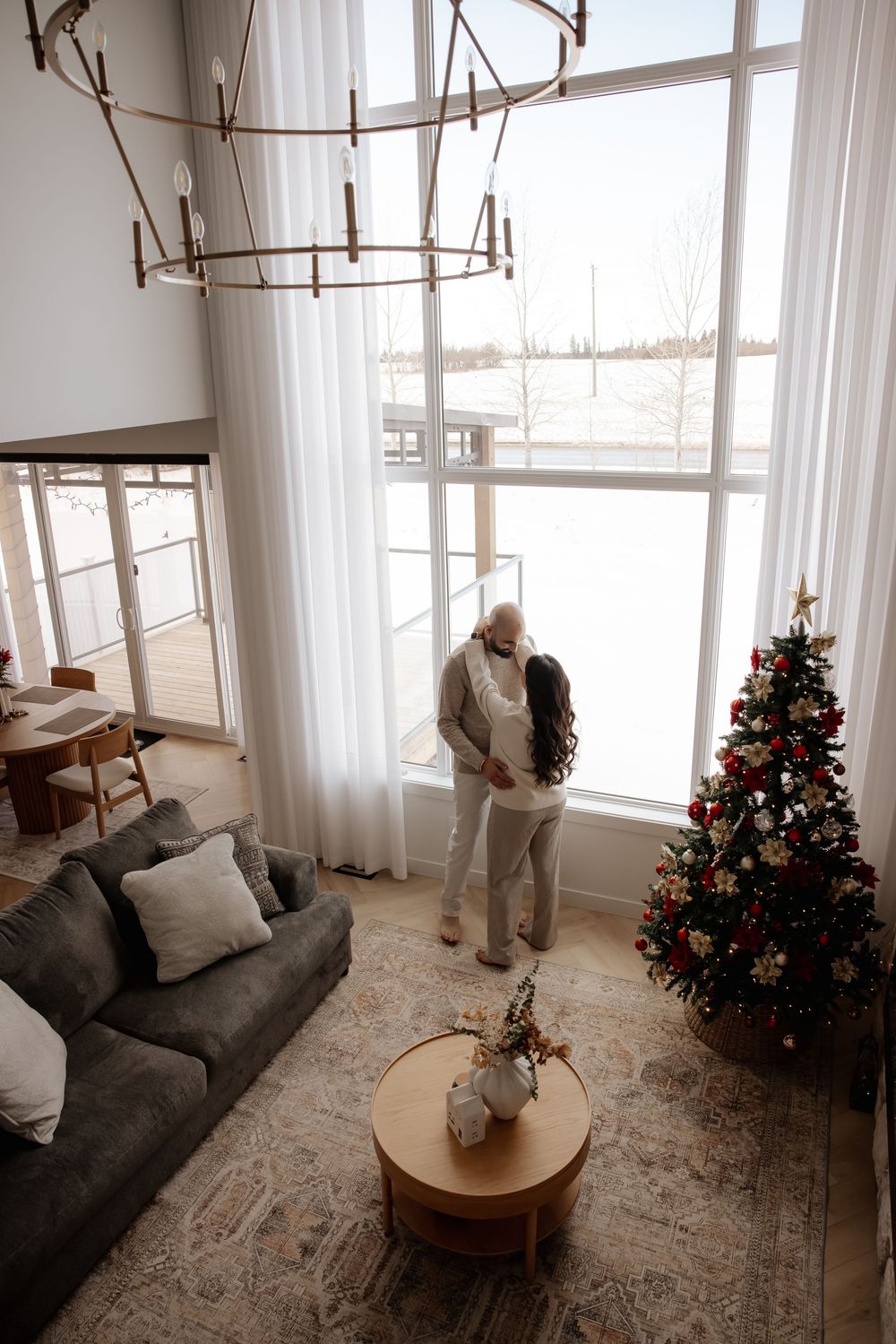 Living room scene with Christmas tree, grey couch, and couple by large windows.