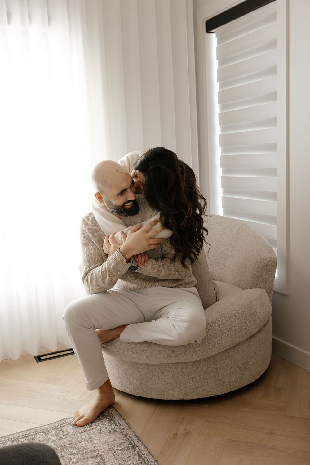 Intimate moments captured on round beige chair near window with white curtains.