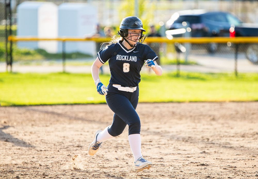 Softball player running on dirt field during game with yellow fence in background.