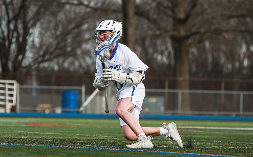 Lacrosse player in white uniform and protective gear kneeling on field.