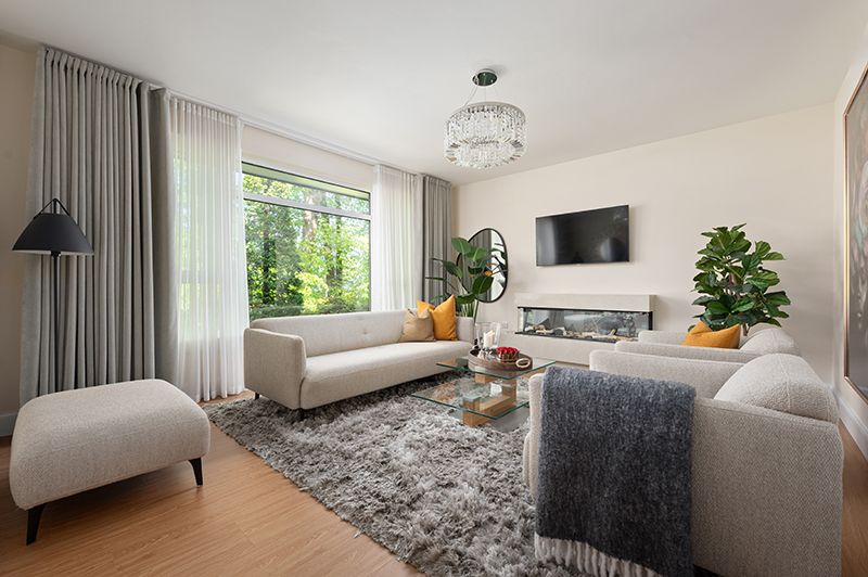 Modern living room with gray sectional sofa, orange accent pillows, hardwood floors and large windows overlooking greenery.