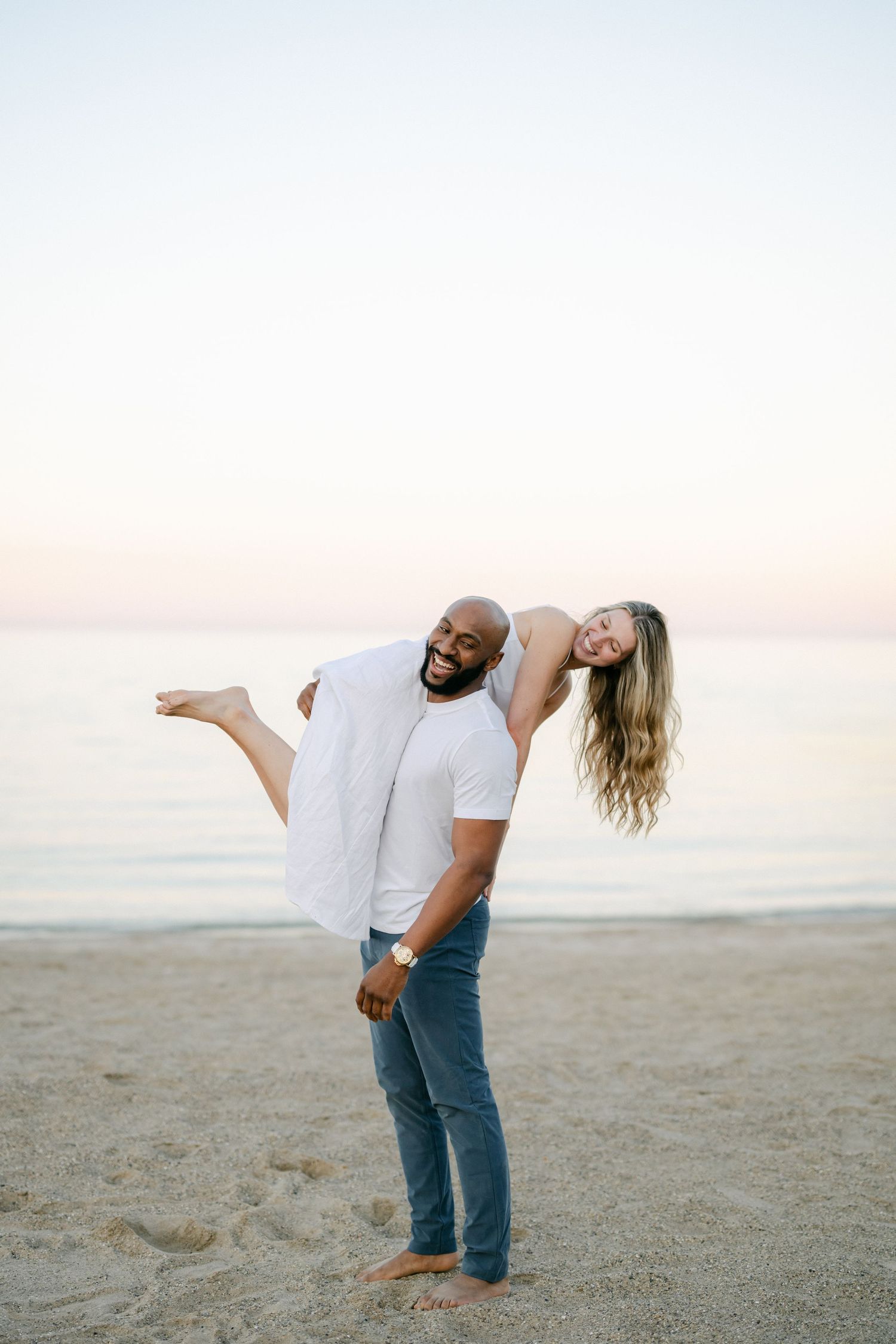 A couple shares a romantic moment on a sandy beach at sunset as they embrace and laugh together. - Lake Forest