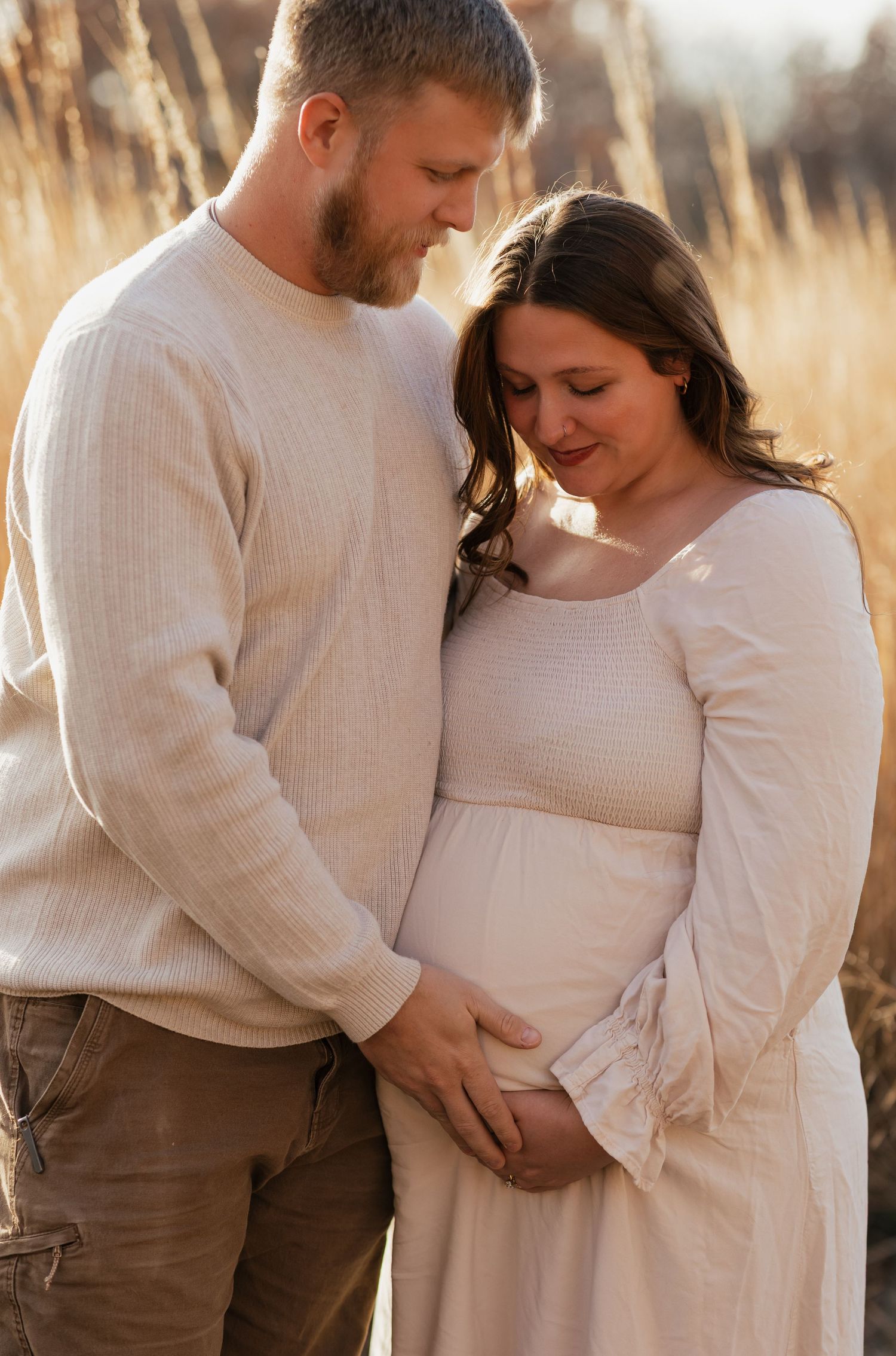 A tender maternity photo of a couple in white sweaters embracing in a sunlit field of tall grass.
