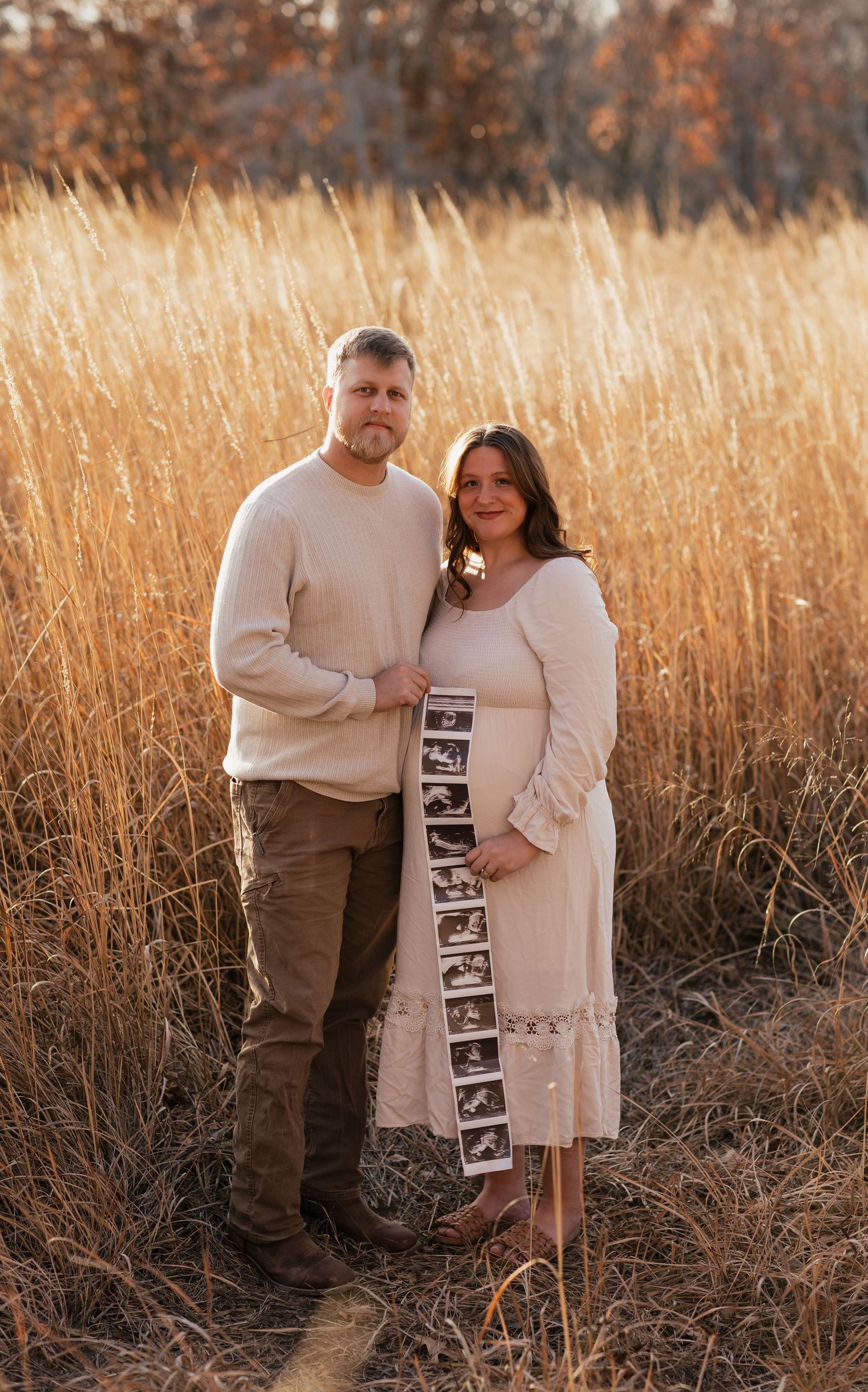 Expectant couple in cream sweaters pose with ultrasound strip in golden autumn field.