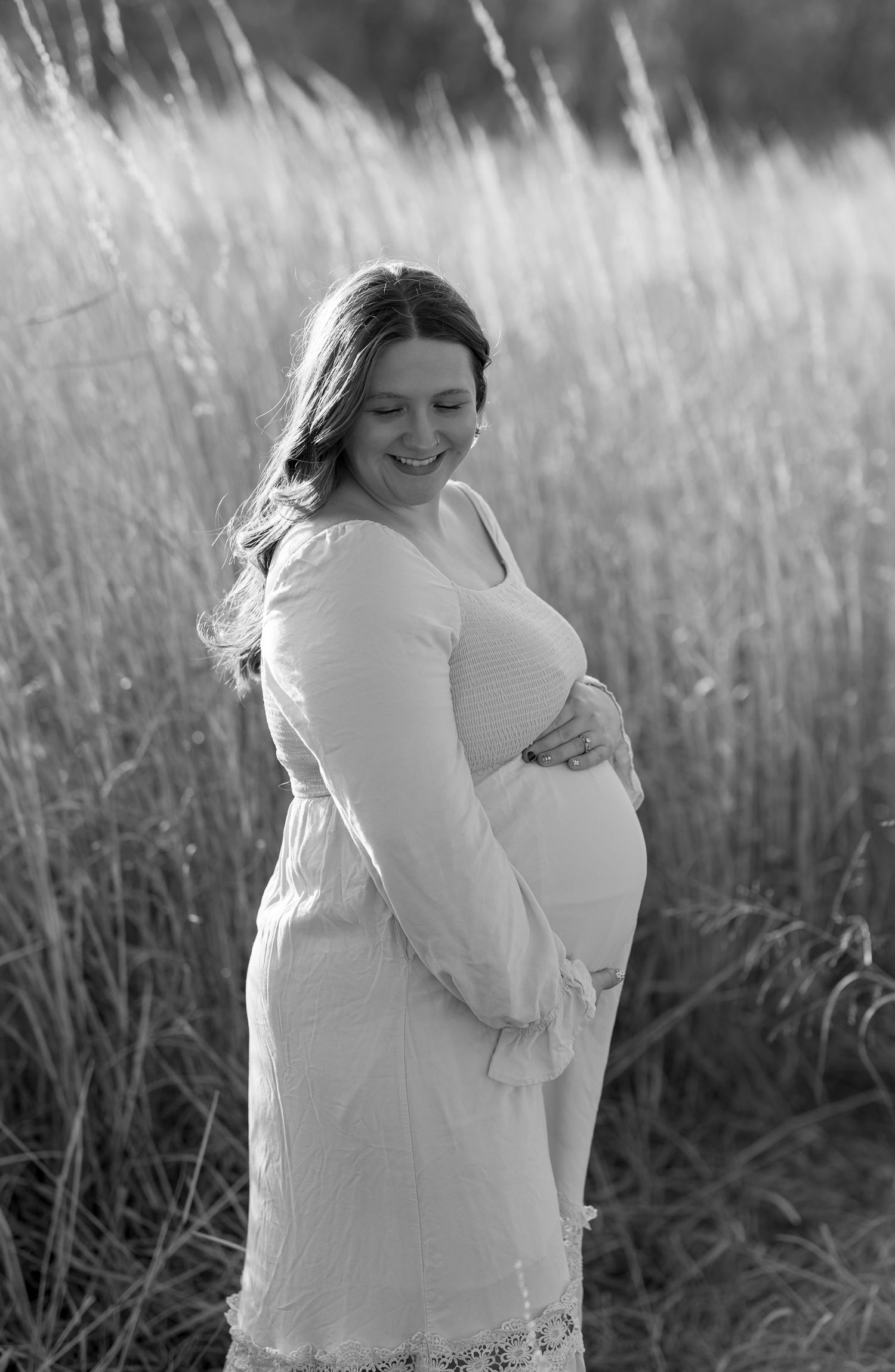 Black and white side profile of expectant mother in white dress standing among tall grass.