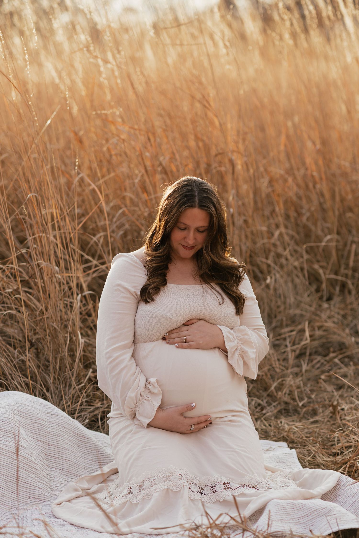 Maternity portrait of woman in white dress sitting among golden autumn grass at sunset.