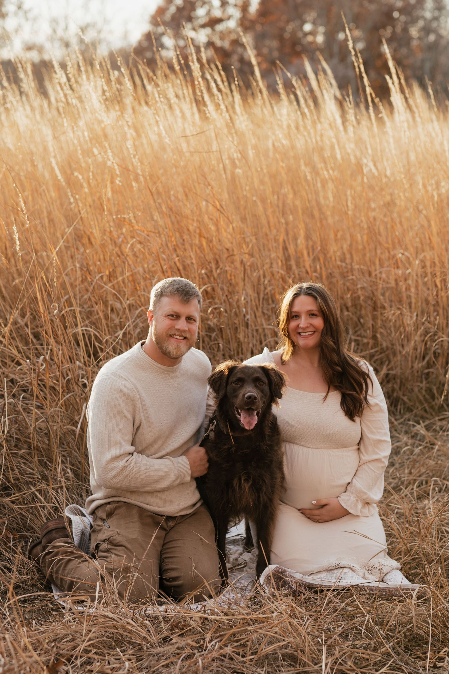 Maternity photo featuring couple with their black dog in a golden field at sunset.