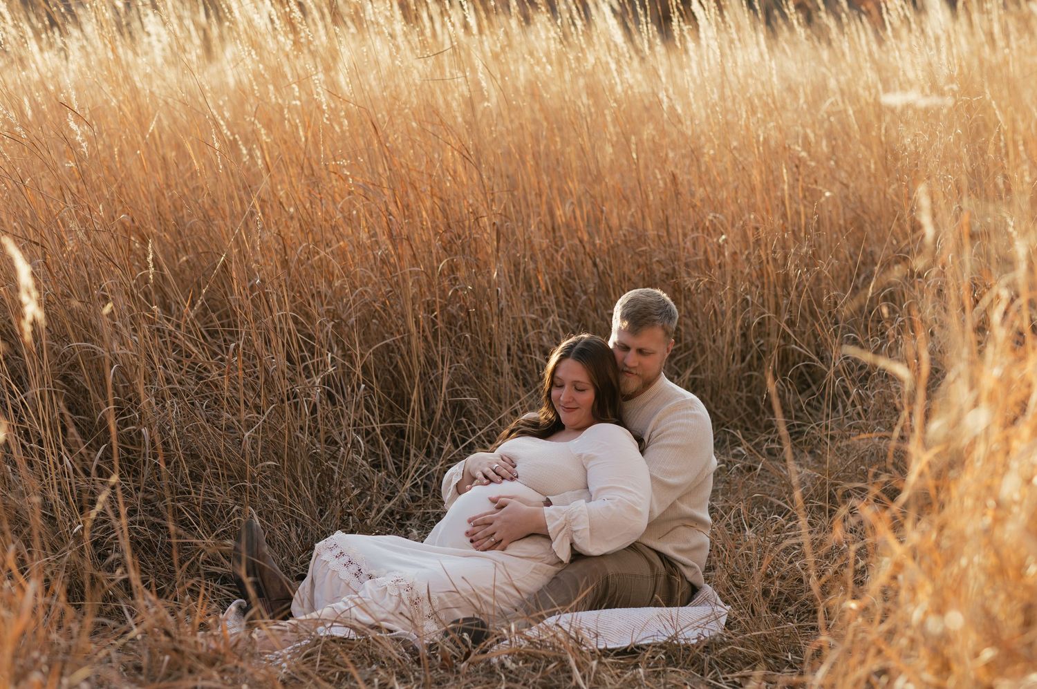 Intimate maternity portrait of couple sitting together in golden field at sunset.