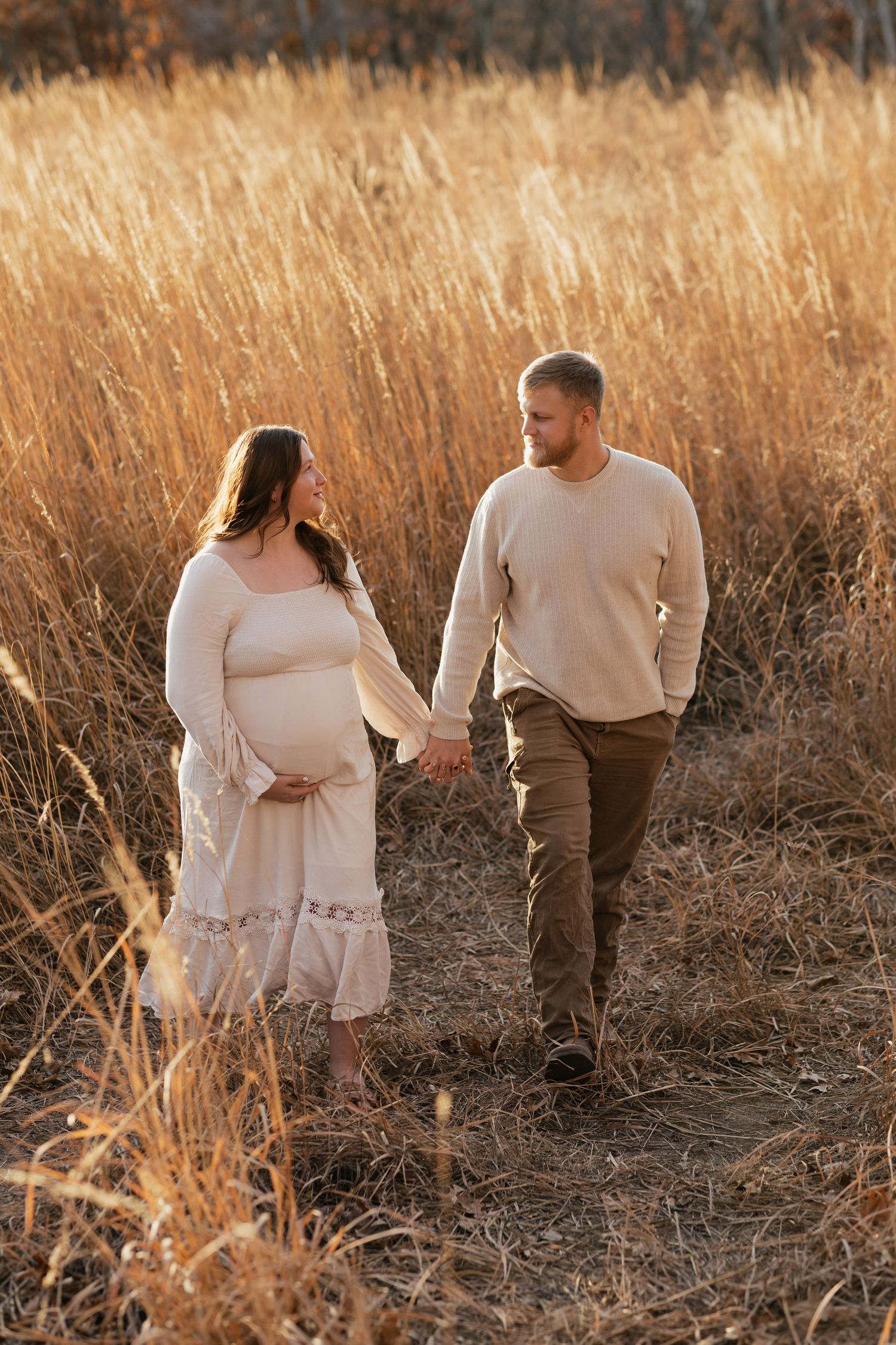 Expecting couple walks hand in hand through tall golden grass at sunset wearing coordinating cream outfits.