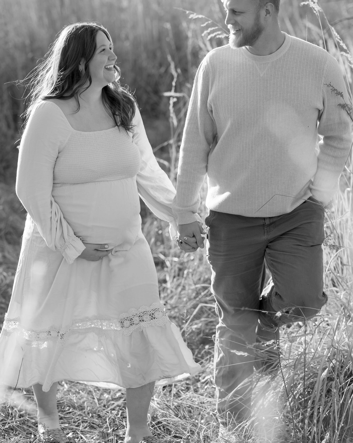 Black and white portrait of a couple sharing a joyful moment among tall grasses during a maternity shoot.