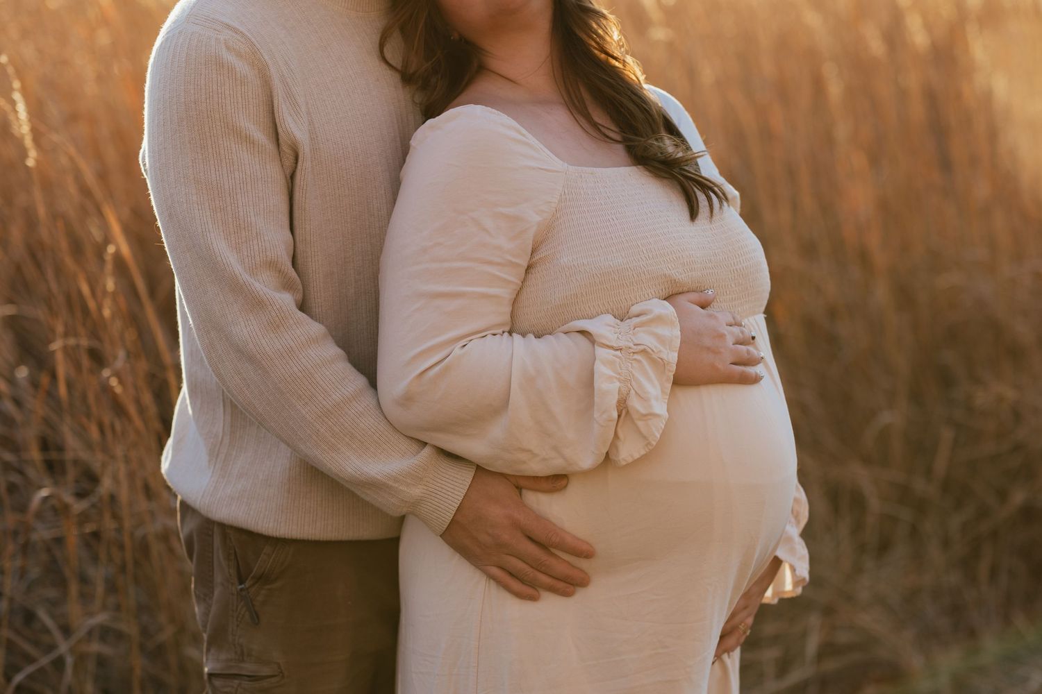 Intimate side view of expecting parents embracing in warm sunset light among tall grasses.