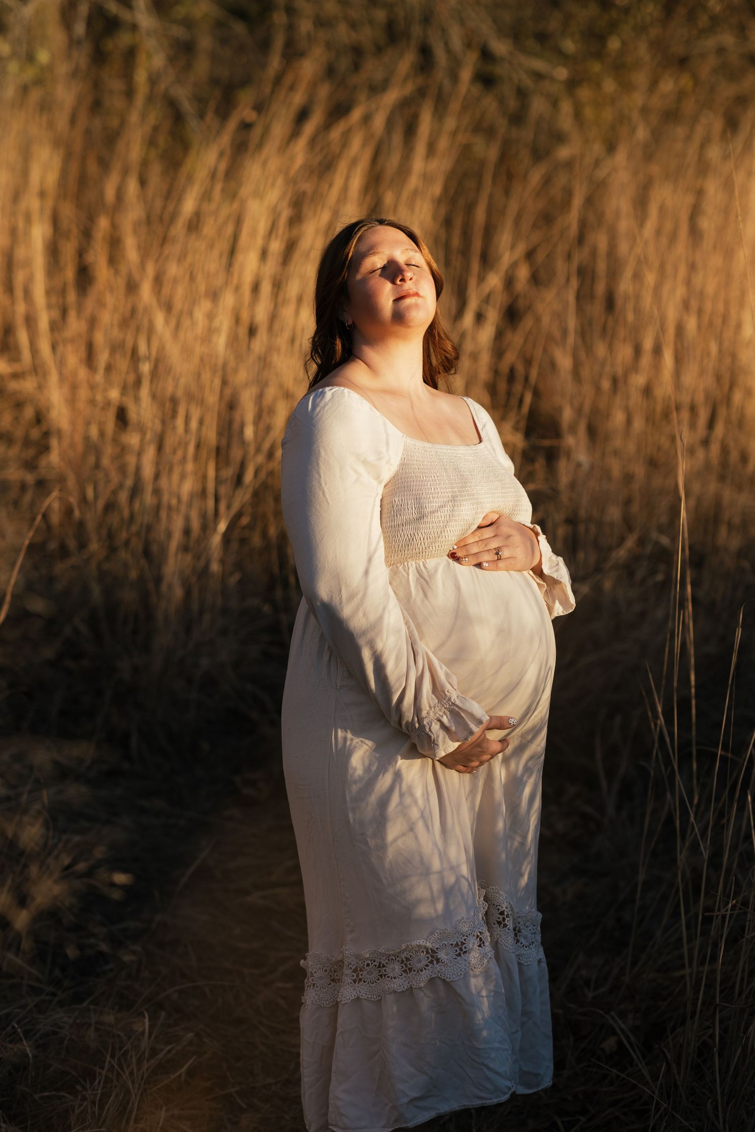 Ethereal maternity portrait in a white dress against golden grass during sunset.