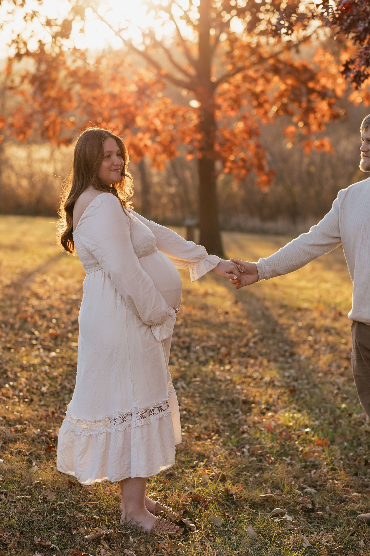 Dreamy autumn maternity photo with couple holding hands in golden sunset light.