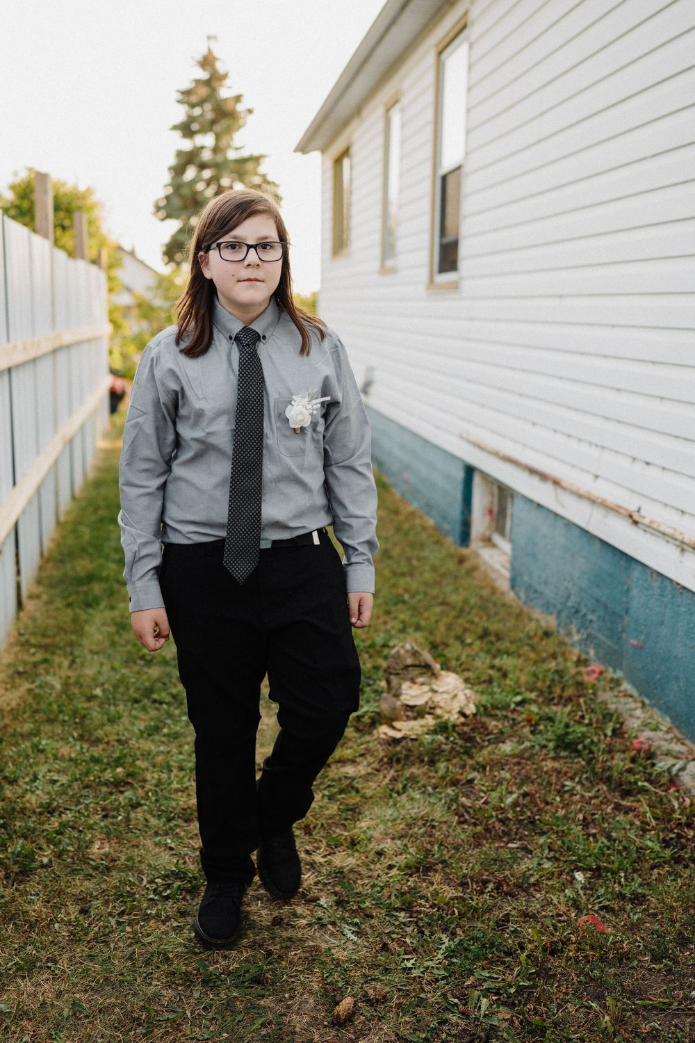 Young boy in gray dress shirt, black tie, and slacks walks outdoors near a white house during a wedding.