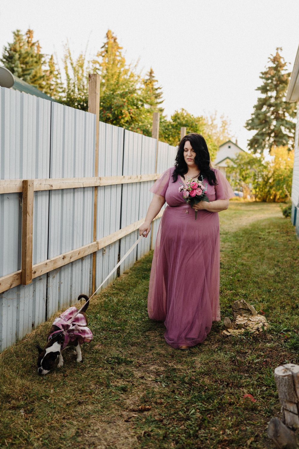 Plus-size bridesmaid in mauve tulle gown holds pink bouquet while walking a dog dressed in pink ribbon outdoors.